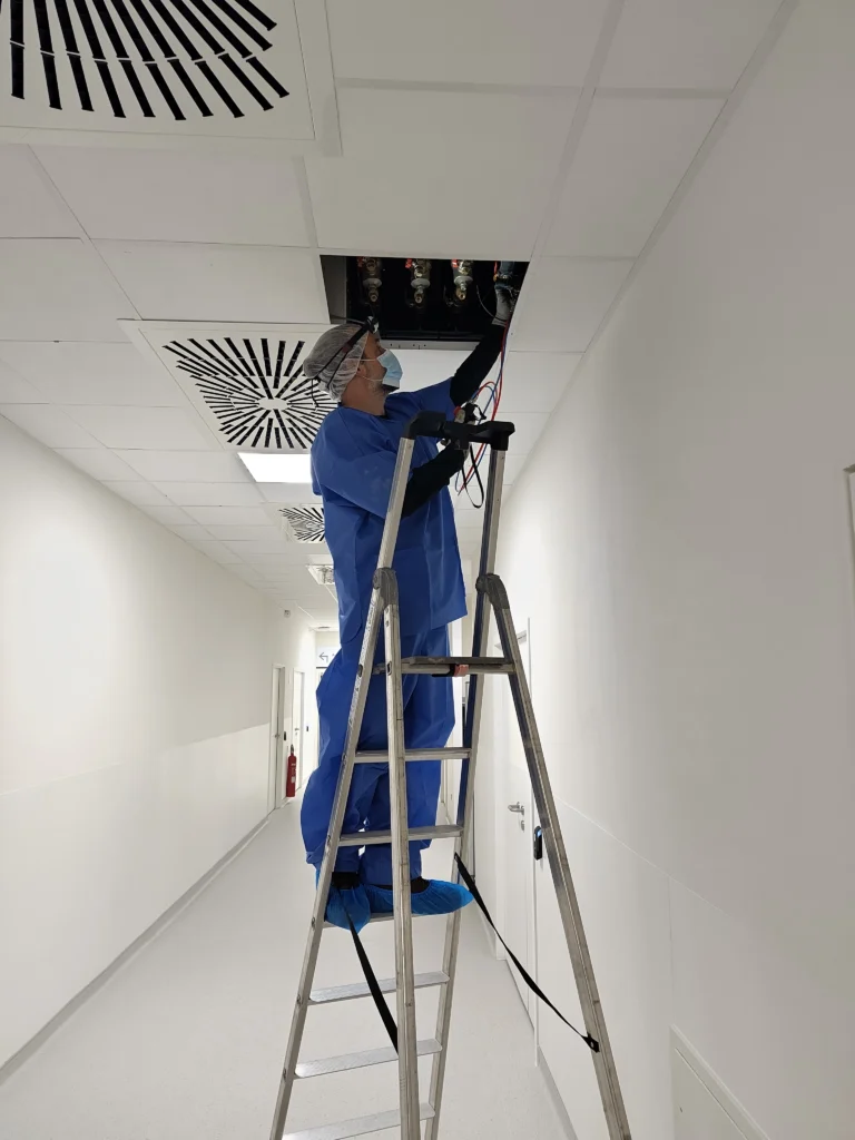 Technicien en tenue stérile effectuant des mesures sur le réseau ECS depuis un escabeau, dans les couloirs des blocs opératoires du nouvel hôpital de Reims